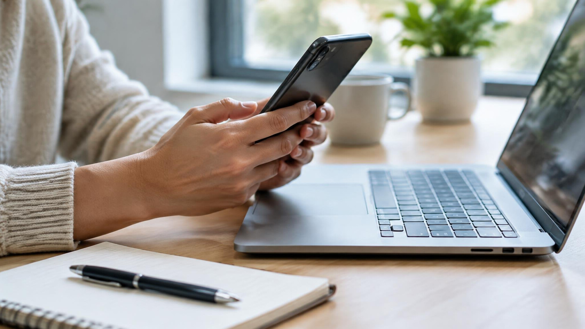 Hands holding a smartphone above an open laptop on a wooden desk